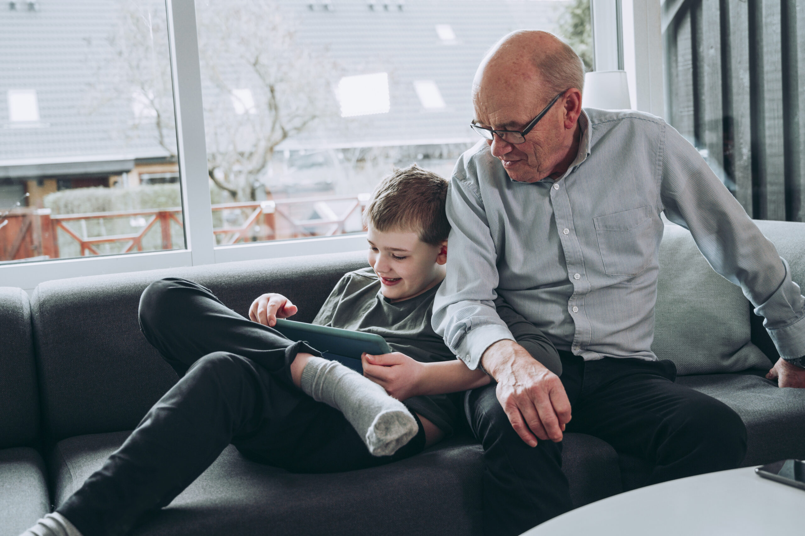 Grandfather and grandson enjoy spending time together playing with tablet at home Grandfather and grandchild sit in a sofa in a living room. The boy holds on to a digital tablet, and they both look at the screen.