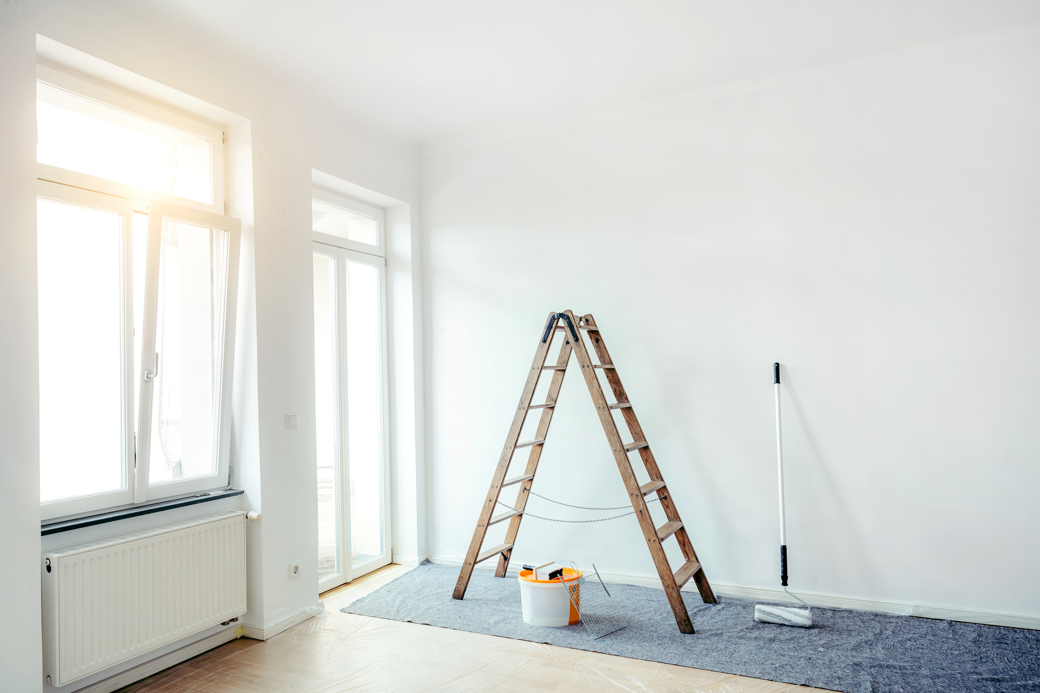 Renovate living room with color bucket and ladder Empty apartment after renovation. Painting in a white room with windows with a stepladder, paint roller and paints on the floor.
